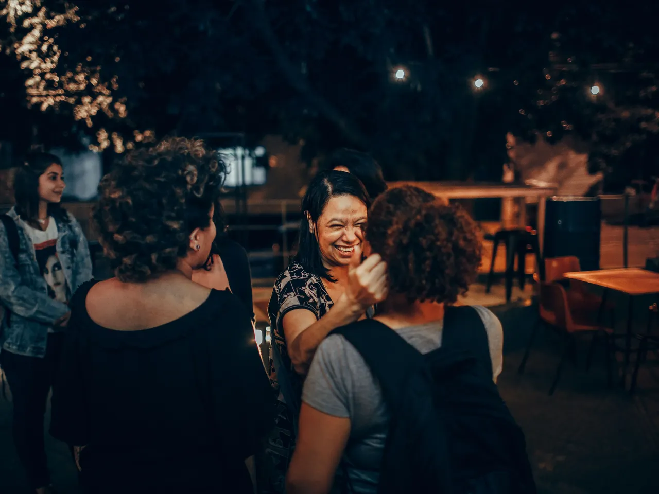 Friends greeting each other at night in the courtyard of an outdoor cafe, under string lights, with empty tables behind them.