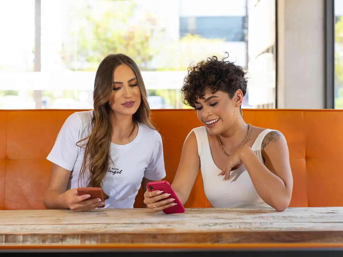 Two friends sit in a café and choose together on their phones.