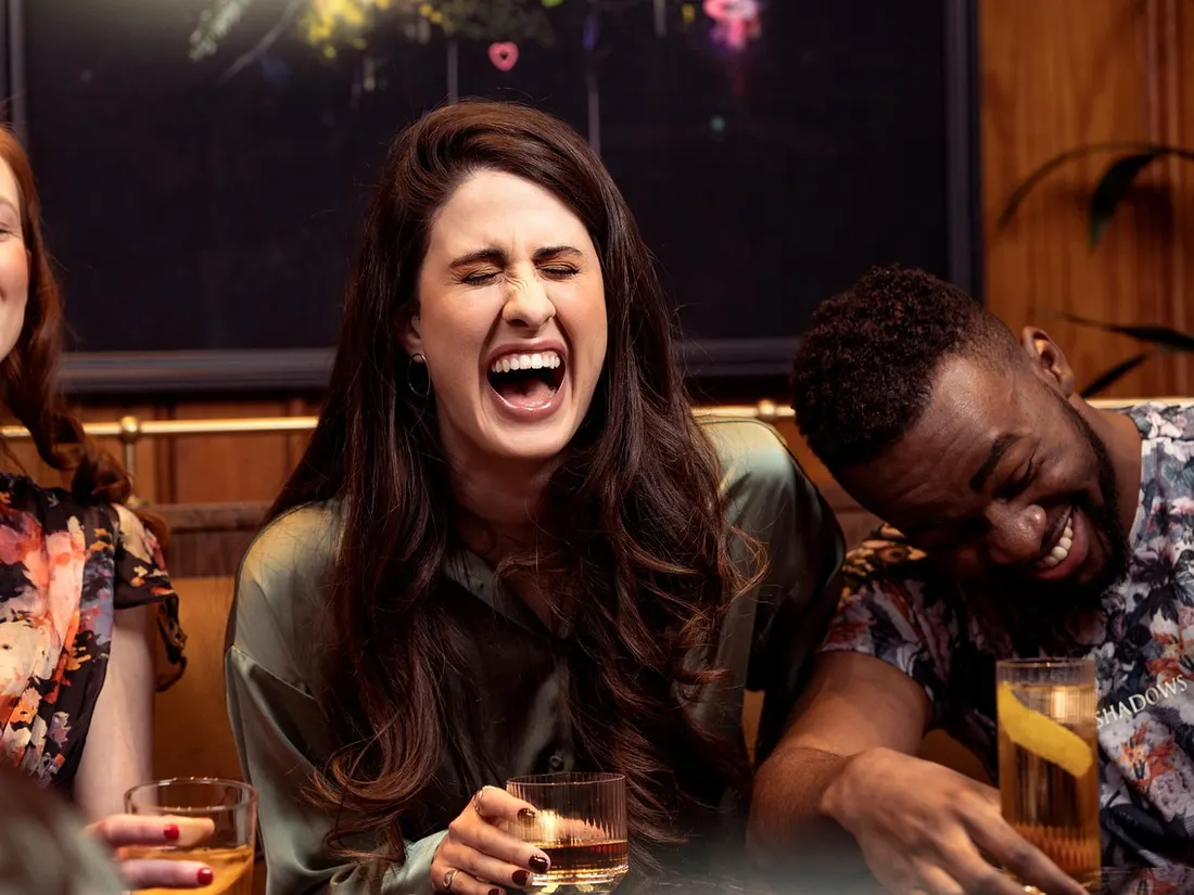Friends laugh together at a table in a warmly lit bar.