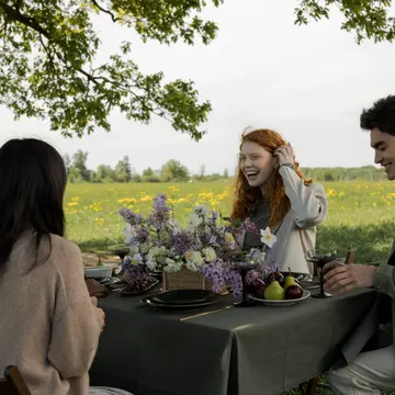 Four friends at a table under a tree in a meadow, laughing over flowers, fruit and glasses.